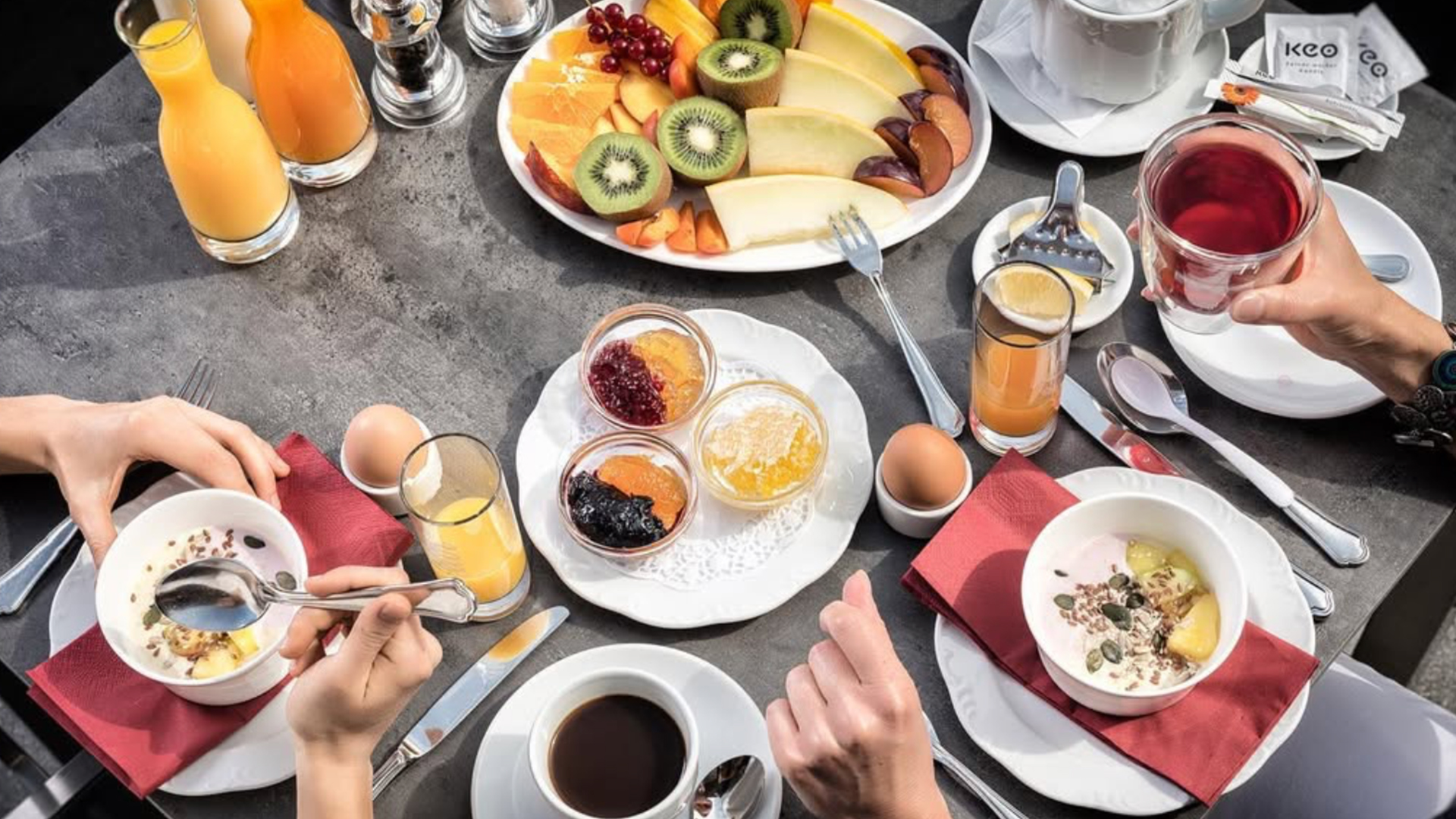 Drei Personen genießen ein Frühstücksbuffet mit Saft, Obst, Kaffee, weichgekochten Eiern, Joghurt mit Müsli und kleinen Gläsern Marmelade, alles auf einem grauen Tisch mit weißen Tellern und roten Servietten.