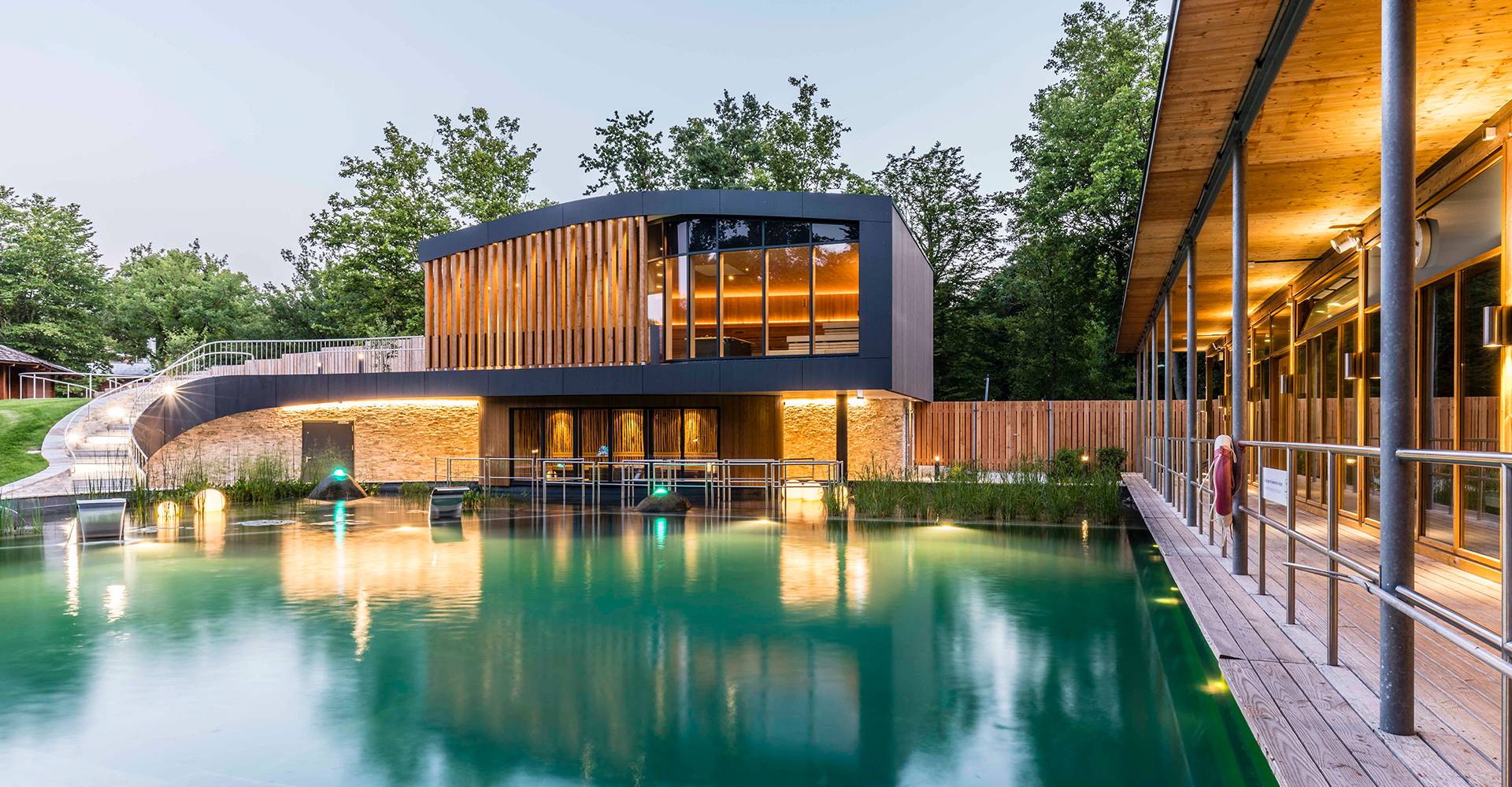 Ein modernes Gebäude mit Holzakzenten blickt auf einen ruhigen, grün gefärbten Teich, der von Bäumen umgeben ist. Warme Lichter beleuchten das Gebäude und spiegeln sich im Wasser, wodurch eine friedliche, einladende Atmosphäre entsteht.