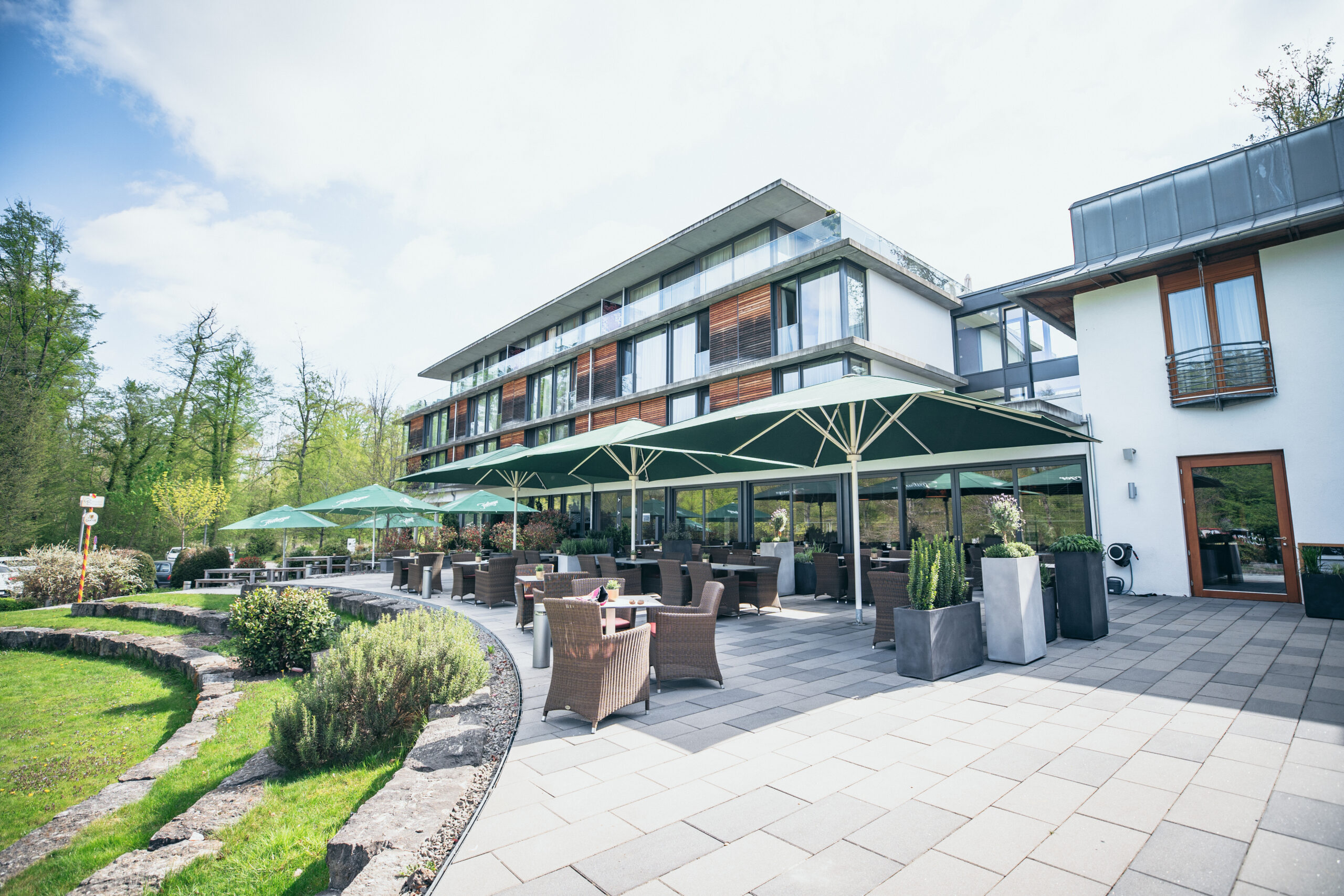 Modern hotel with large windows and balconies, featuring an outdoor patio area with wicker chairs, tables, and green umbrellas. Lush greenery and trees surround the building under a partly cloudy sky.