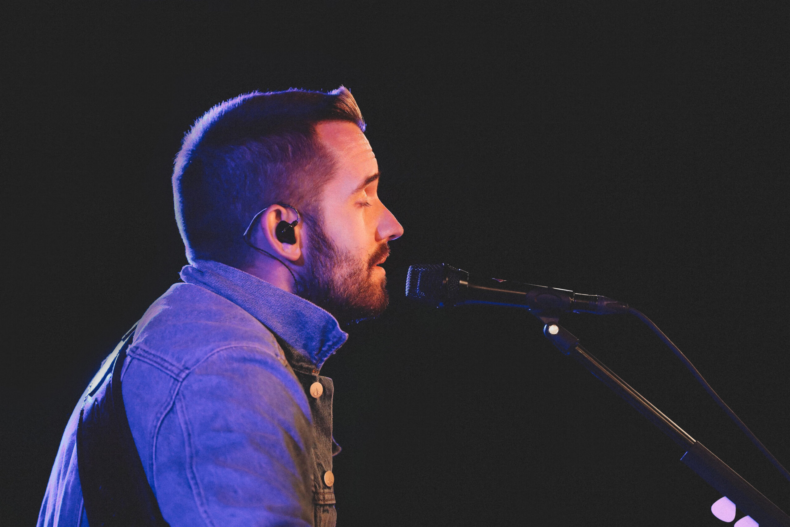 Ein bärtiger Mann in Jeansjacke und mit In-Ear-Monitor singt auf der Bühne bei dramatischer Beleuchtung in ein Mikrofon.