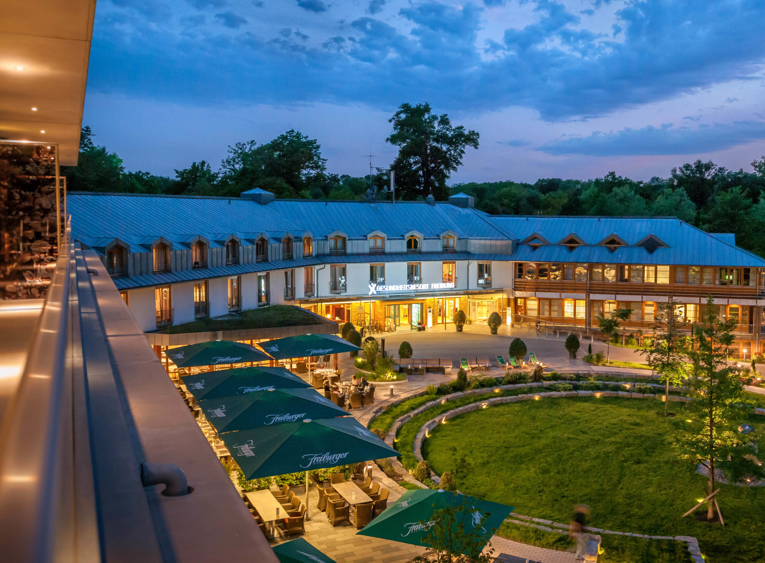 Ein großes, modernes Hotel mit Sitzgelegenheiten im Freien und grünen Sonnenschirmen umgibt eine runde Rasenfläche in der Abenddämmerung, während das Gebäude von Lichtern beleuchtet wird und der blaue Himmel mit Wolken bedeckt ist.