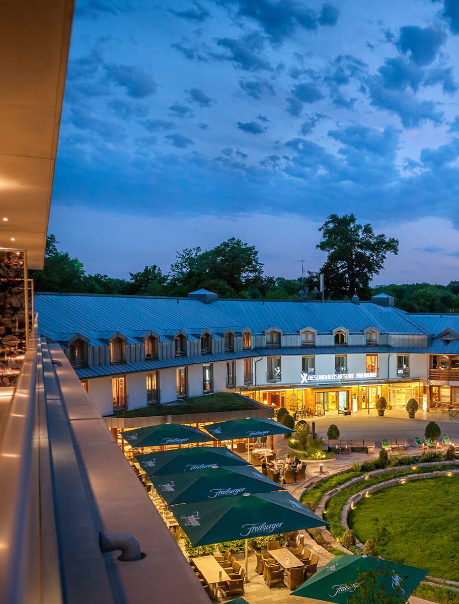 Ein Blick auf einen Hotelhof in der Abenddämmerung, mit beleuchteten Fenstern, Sitzgelegenheiten im Freien unter grünen Sonnenschirmen und einem üppigen grünen Rasen, umgeben von Bäumen unter einem teilweise bewölkten Abendhimmel.