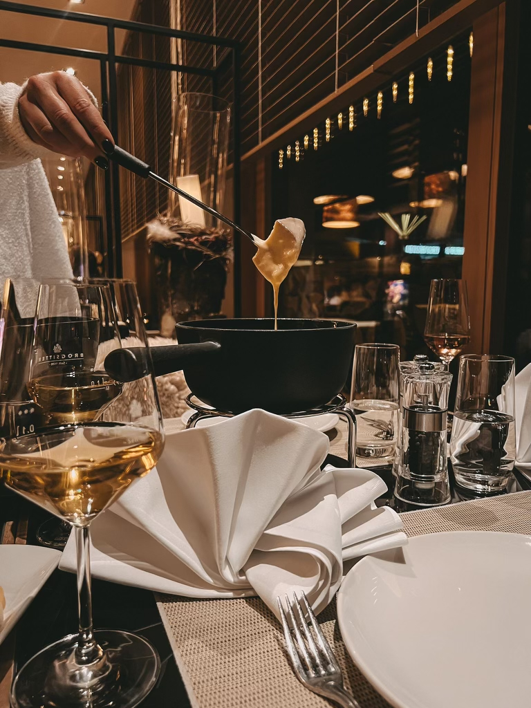 Eine Person taucht Brot in einen Topf mit geschmolzenem Käsefondue an einem mit Weingläsern, Tellern und Servietten gedeckten Restauranttisch, der eine gemütliche Atmosphäre schafft.