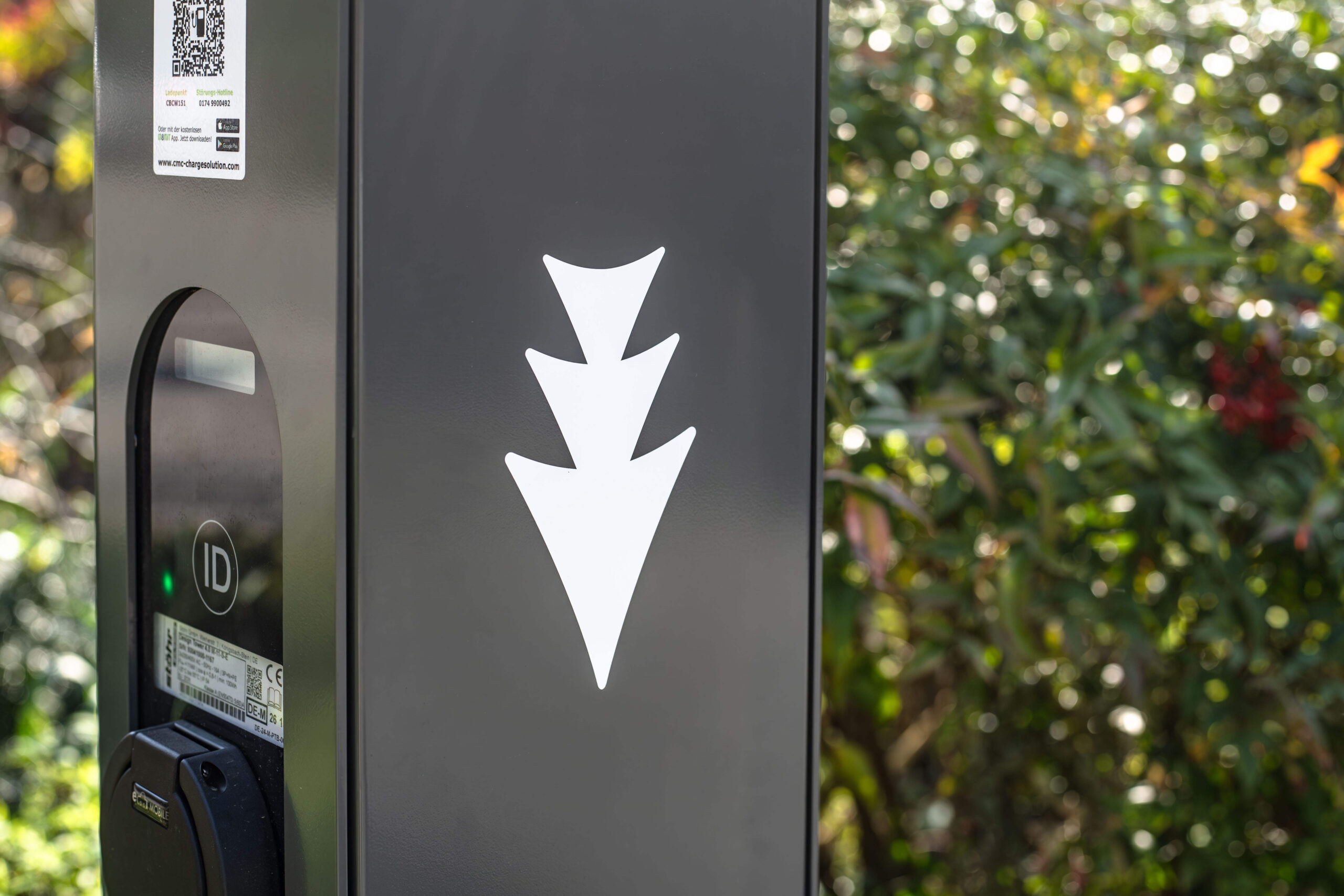 Eine Nahaufnahme einer schwarzen Ladestation für Elektrofahrzeuge mit einem weißen, nach unten zeigenden Dreipfeilsymbol, umgeben von Grün und Sonnenlicht im Hintergrund.