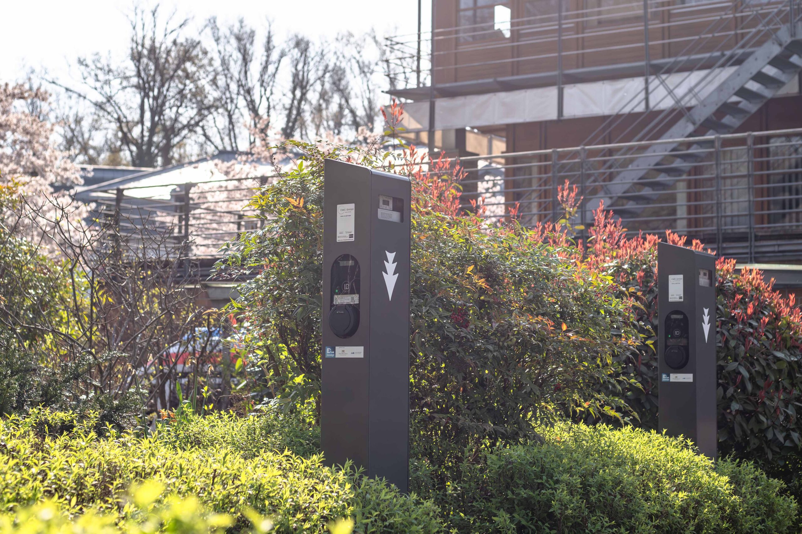 Zwei Ladestationen für Elektrofahrzeuge stehen zwischen grünen Büschen und rotblättrigen Pflanzen. Im Hintergrund sind an einem sonnigen Tag ein modernes Gebäude und eine Metalltreppe zu sehen.