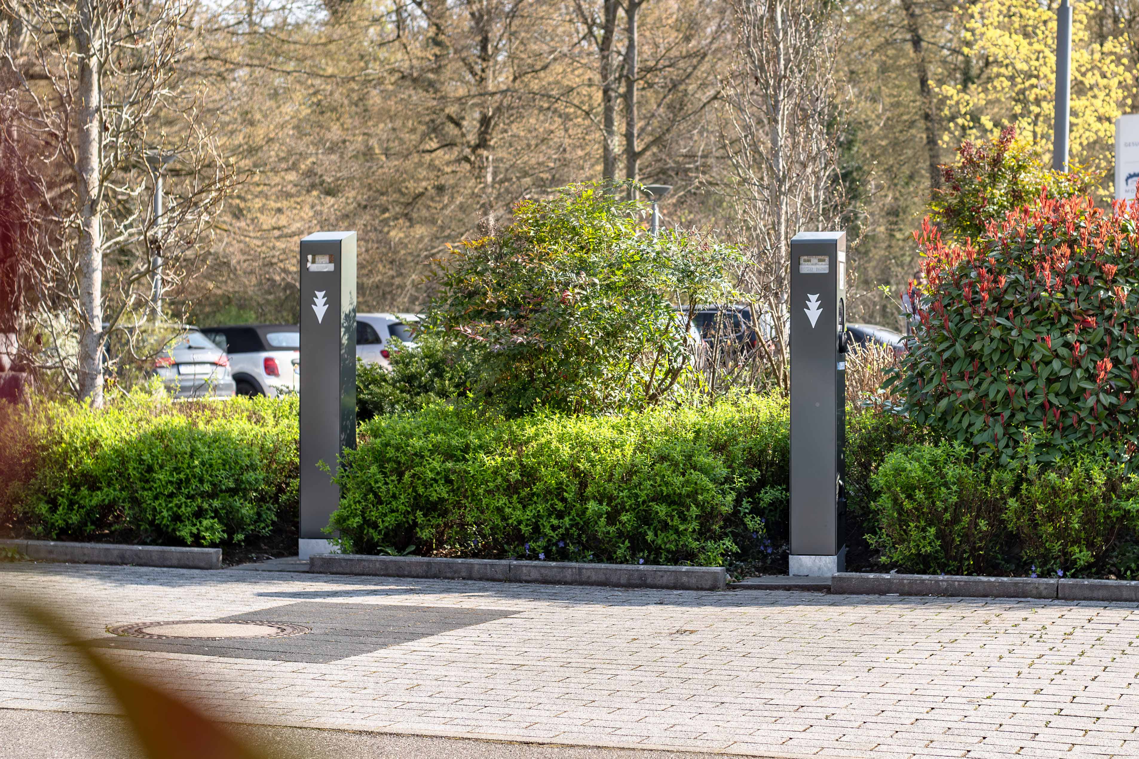 Zwei Ladestationen für Elektrofahrzeuge stehen auf einem Parkplatz, umgeben von grünen Büschen und Bäumen. Im Hintergrund sind an einem sonnigen Tag geparkte Autos zu sehen.
