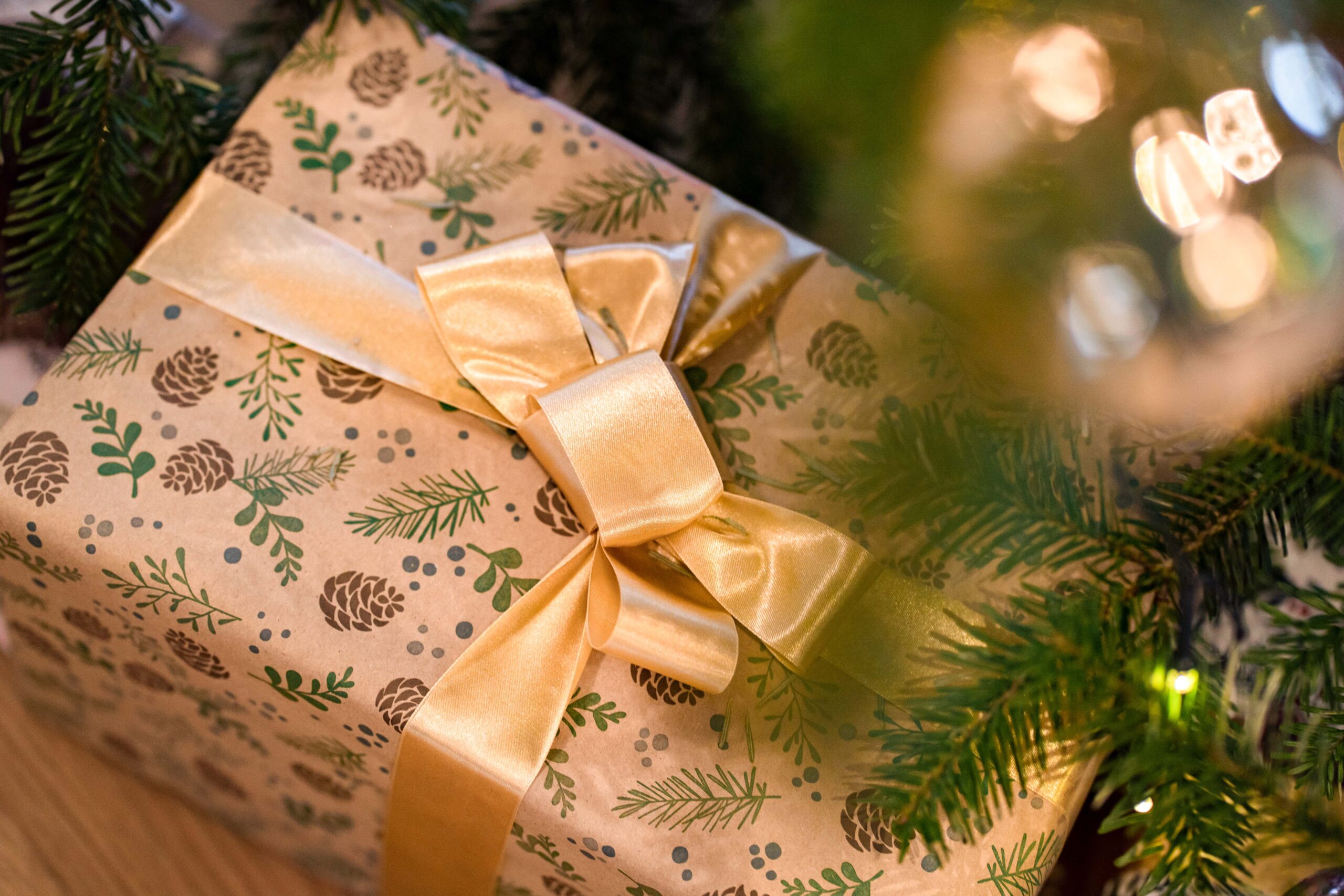 Ein eingepacktes Geschenk liegt unter einem Weihnachtsbaum. Auf dem Papier sind Tannenzapfen und grüne Zweige zu sehen, und die Schachtel ist mit einem glänzenden Goldband und einer Schleife verschnürt. Im Vordergrund sind verschwommene Zweige und Lichterketten zu sehen.
