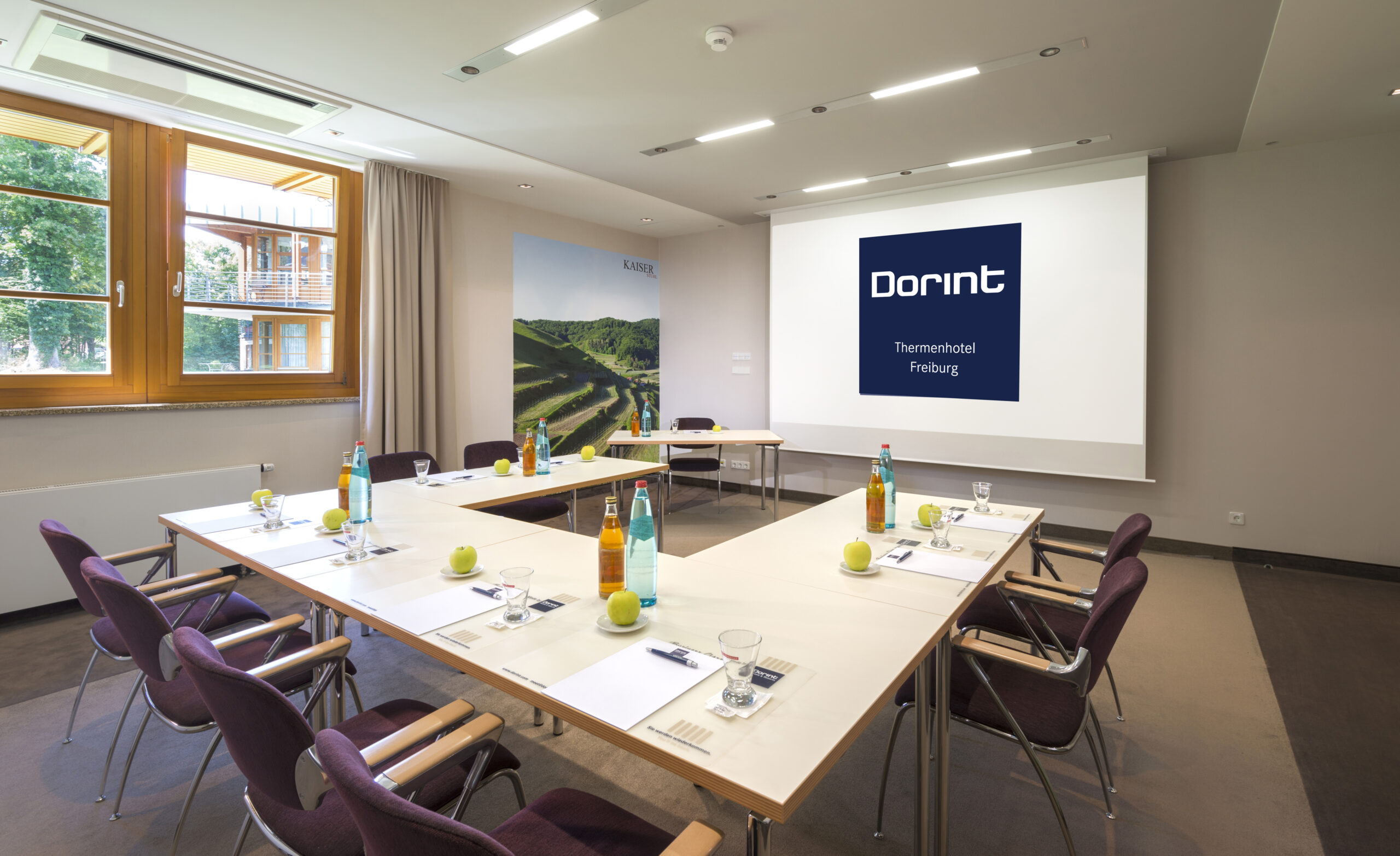A modern conference room with a U-shaped table set for a meeting. Each seat has a pen, notepad, glass, water, and an apple. Bottles of water and juice are on the table. A large screen displays the Dorint logo.