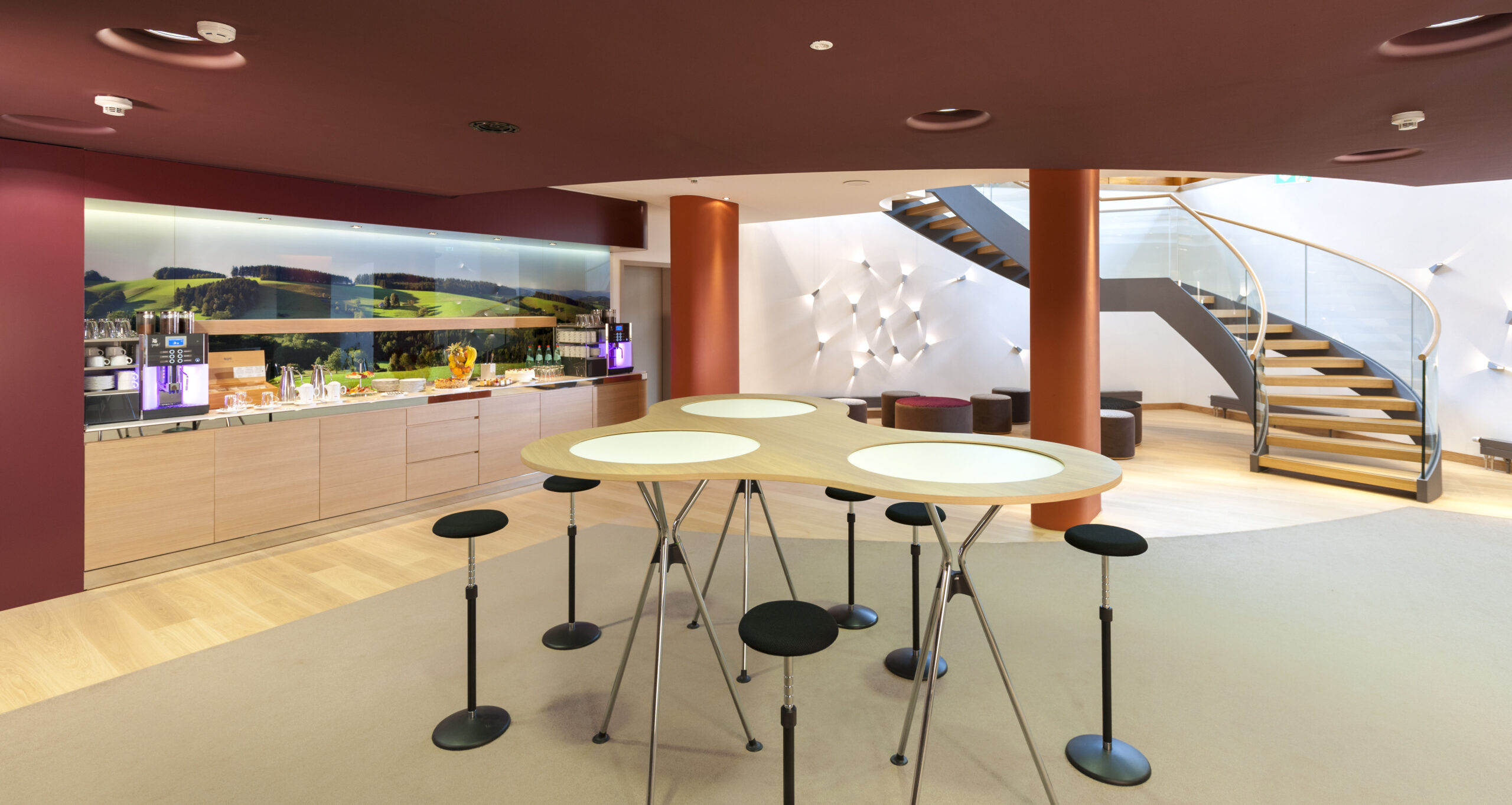 Moderner Lounge-Bereich mit Stehtischen und Barhockern, einem Erfrischungstresen mit Getränken und Snacks, Holzfußboden und einer geschwungenen Treppe mit Glasgeländer, die in eine obere Etage führt. Zeitgemäße Beleuchtung an den Wänden.