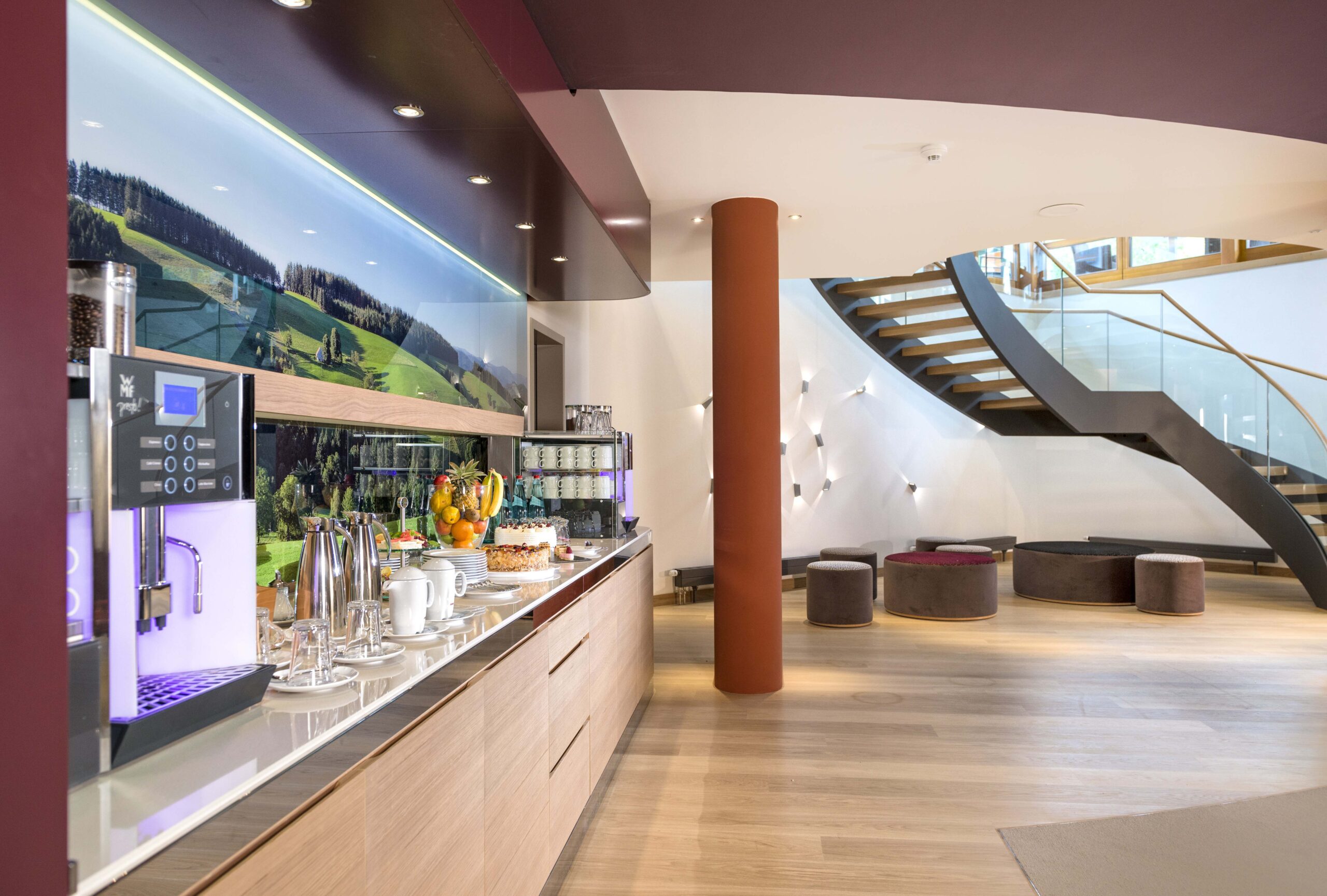 Moderne Hotellobby mit einer Kaffeestation auf der linken Seite, die Getränke, Gebäck und Obst anbietet. Der Bereich verfügt über helle Holzböden, eine geschwungene Treppe und moderne Sitzgelegenheiten mit runden Hockern.