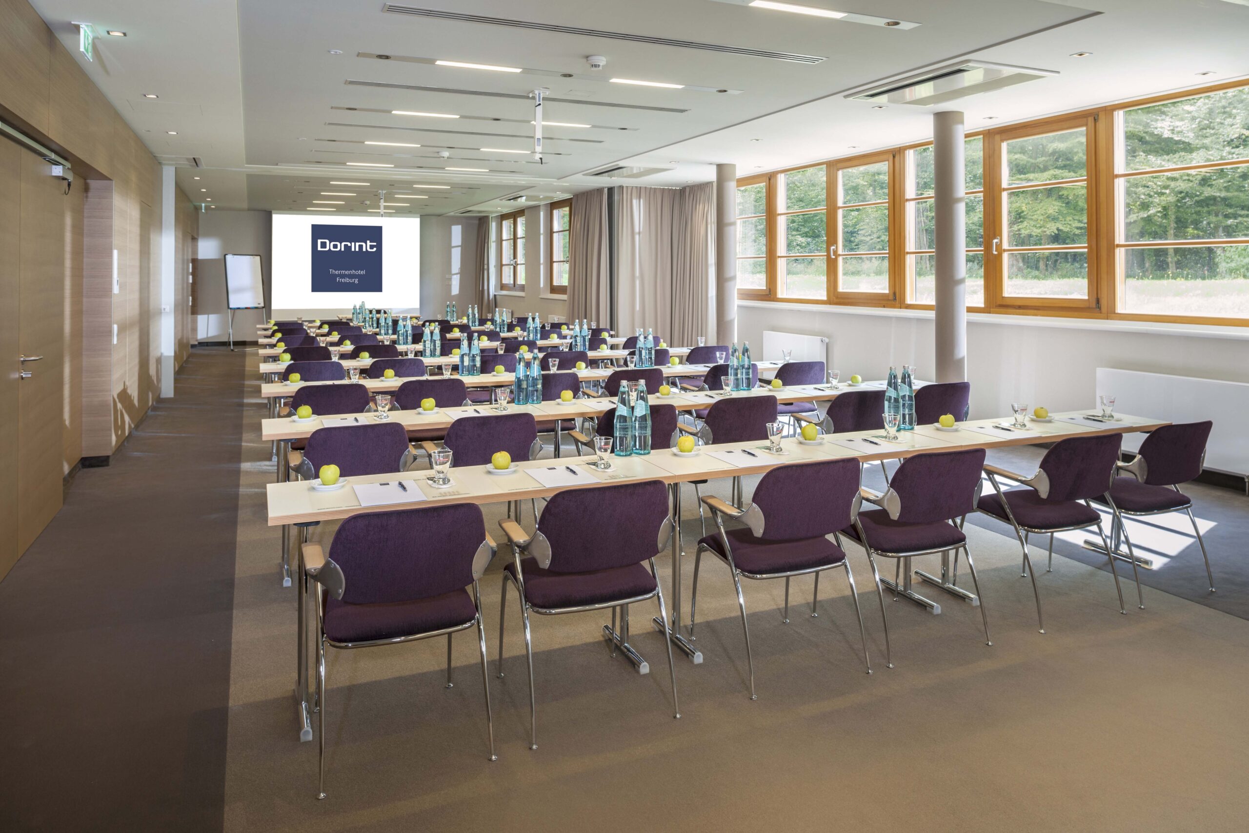 Ein moderner Konferenzraum mit Tischreihen und lilafarbenen Stühlen vor einer Projektionsfläche. Auf jedem Tisch stehen Wasserflaschen, Gläser, Notizblöcke, Stifte und grüne Äpfel. Große Fenster lassen natürliches Licht herein.