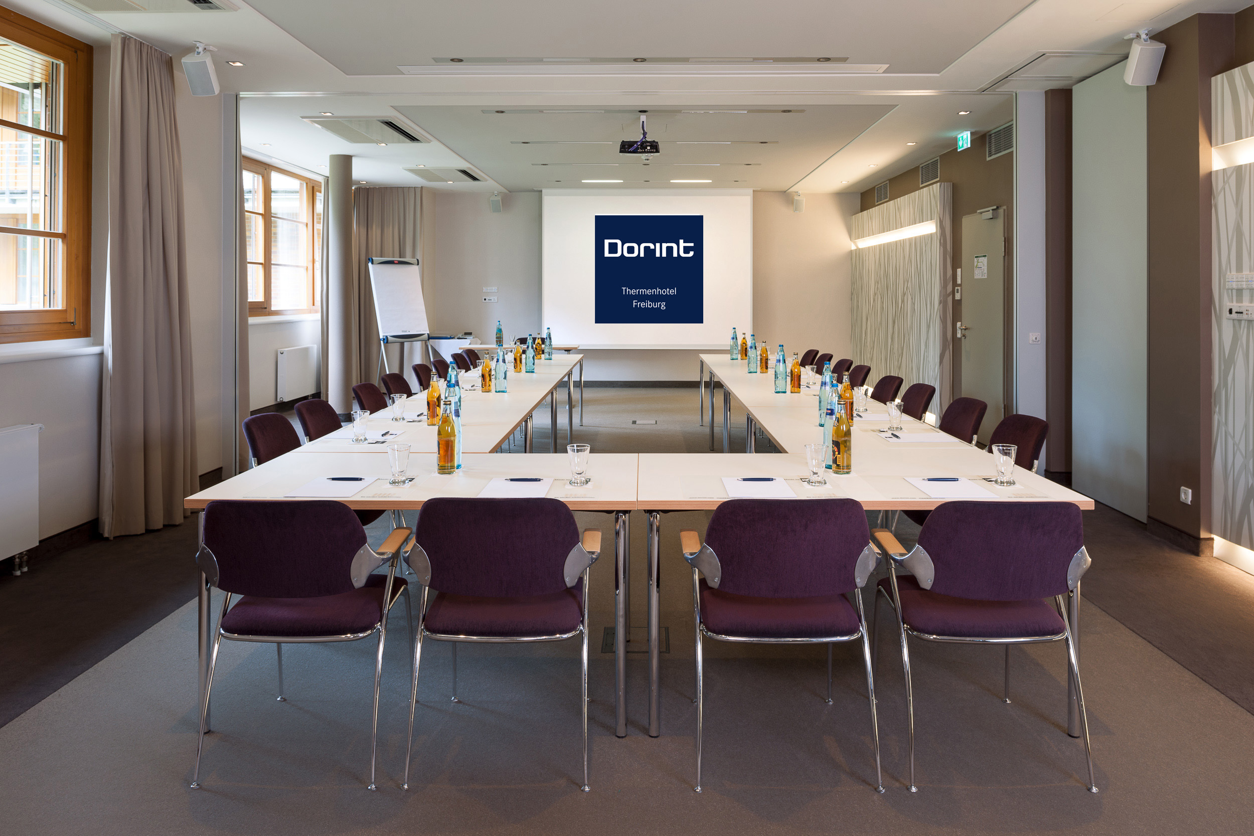 Ein moderner Konferenzraum mit U-förmigen Tischen, lila Stühlen, Getränkeflaschen, Notizblöcken und Stiften. Auf einem Bildschirm an der Stirnseite steht "Dorint Thermenhotel Freiburg". Der Raum ist durch Fenster und Deckenlampen gut beleuchtet.