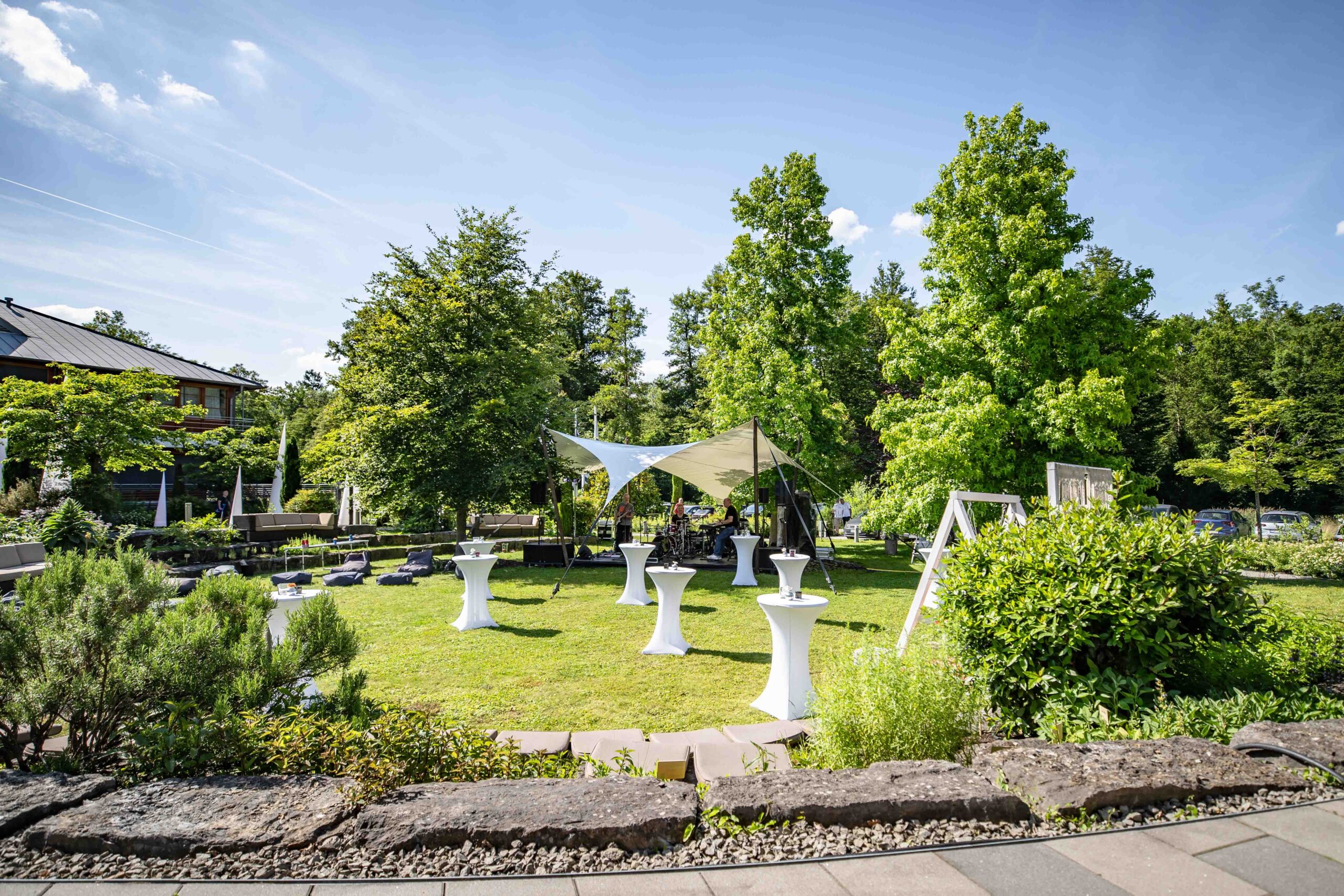A sunny outdoor garden event setup with cocktail tables covered in white cloth, a canopy tent, and lush green trees and bushes surrounding the area. A building is visible on the left.