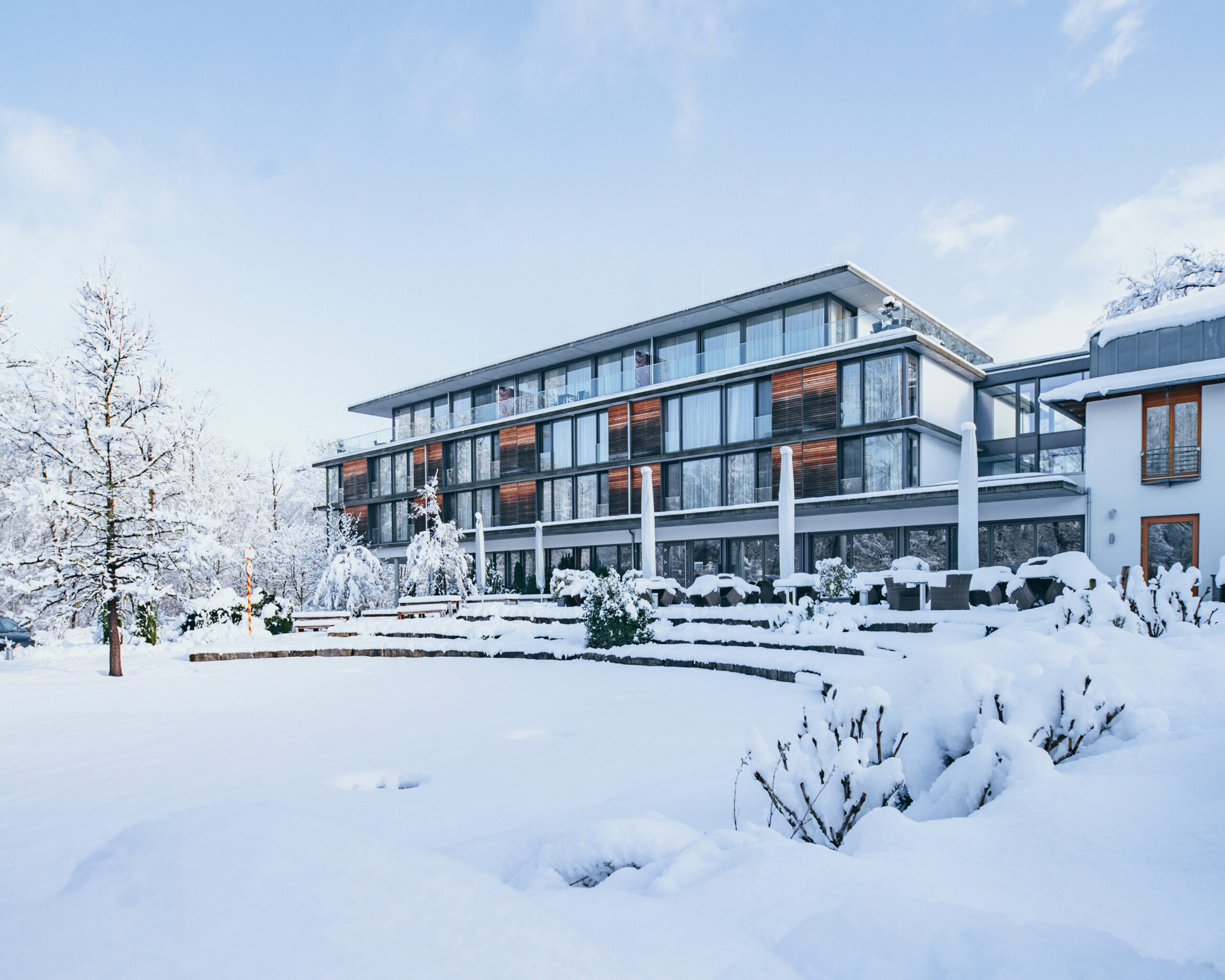 Ein modernes mehrstöckiges Gebäude mit großen Fenstern ist umgeben von schneebedeckten Bäumen, Sträuchern und einer verschneiten Landschaft unter einem teilweise bewölkten Himmel.