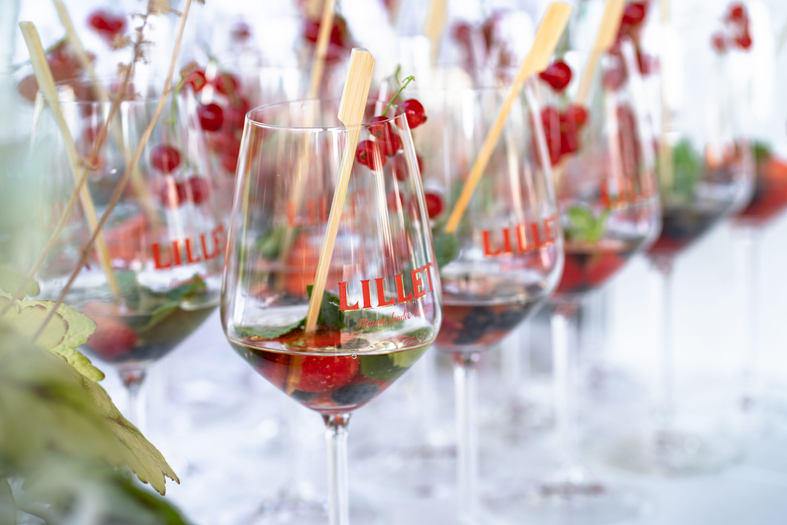Nahaufnahme mehrerer Weingläser mit der Aufschrift Lillet, gefüllt mit einem Cocktail aus Beeren, Kräutern und Eis, garniert mit Johannisbeeren und einem Holzrührer. Die Umgebung wirkt festlich und hell.